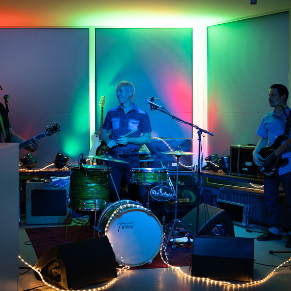Rockband mit drei Musikern auf der Bühne: Gitarrist links, Schlagzeuger in der Mitte und Bassist rechts, beleuchtet von farbigen LED-Panels in Rot, Grün und Blau