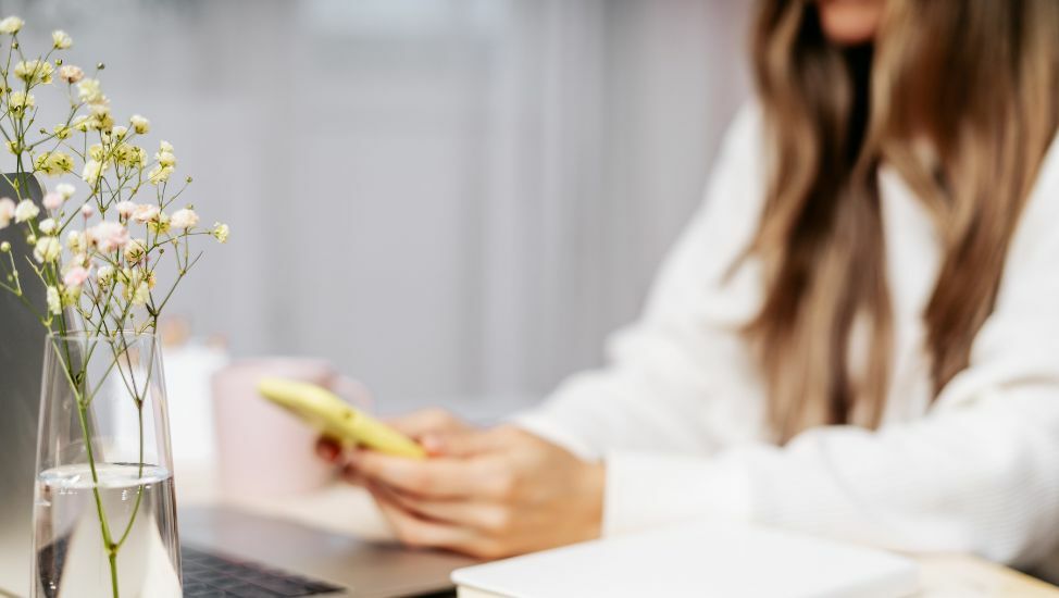Frau mit weißer Bluse und langen Haaren sitzt am Schreibtisch und betrachtet ihr Smartphone in der Hand. Im Vordergrund steht eine  Blume.