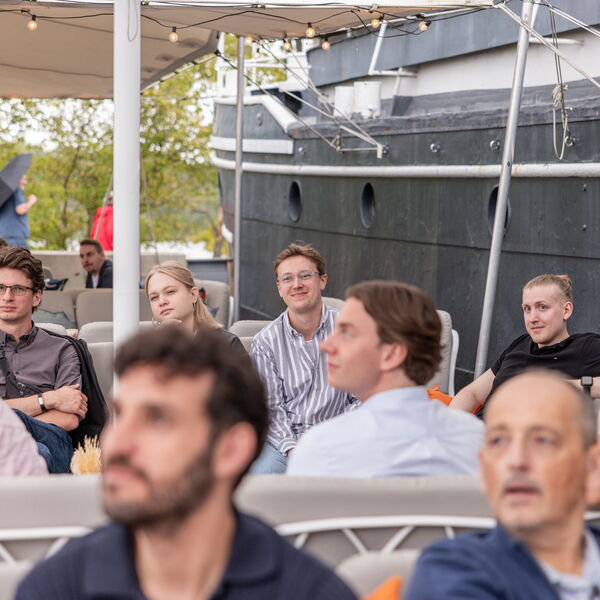 Publikum sitzt unter einem Zelt mit Lichterketten und hört konzentriert zu, im Hintergrund ein Schiff und grüne Bäume