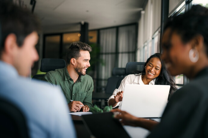 Vier Personen in einem modernen Büro sitzen an einem Tisch und arbeiten gemeinsam an Laptops, zwei von ihnen lächeln sich an