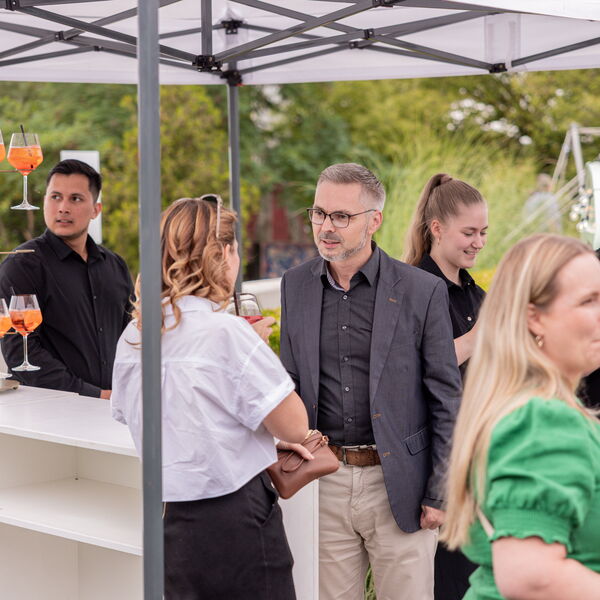 Aperol-Bar unter Zelt mit Barkeeper hinter Tresen, mehreren Gästen und Gläsern gefüllt mit orangenem Cocktail im Grünen