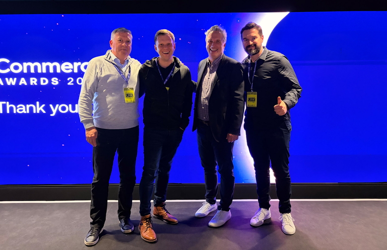Vier Männer mit Event-Badges stehen lächelnd vor einer blauen Bühne bei einer Commerce Awards Veranstaltung.