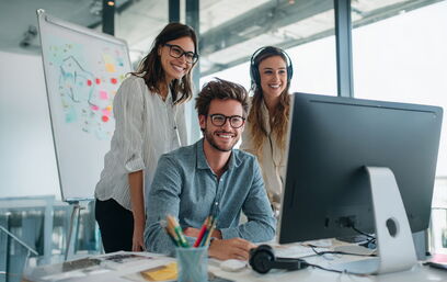 Drei junge Leute arbeiten gemeinsam lächelnd am Computer im modernen Büro